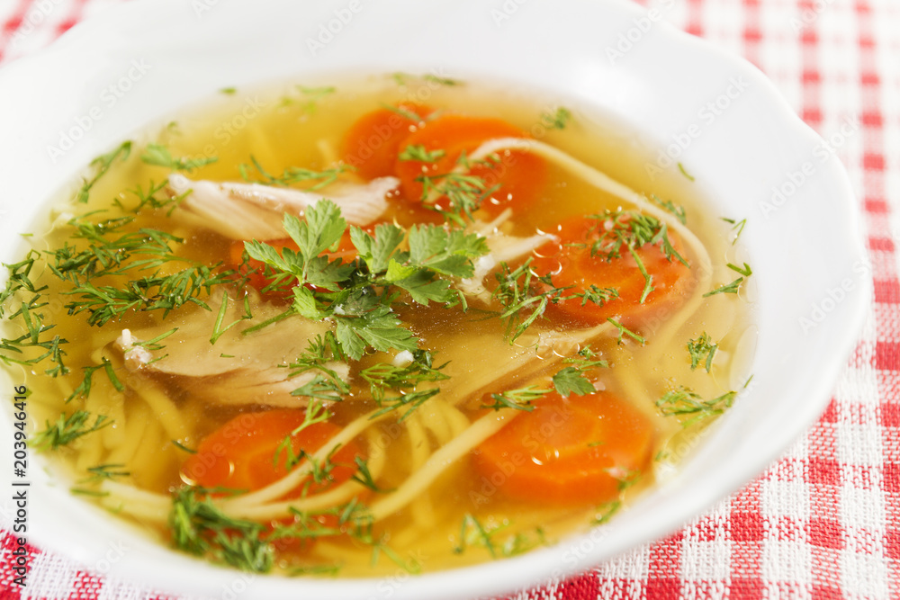Chicken soup or broth with noodels, chicken meat pieces , carrot slices and herbs in white bowl.