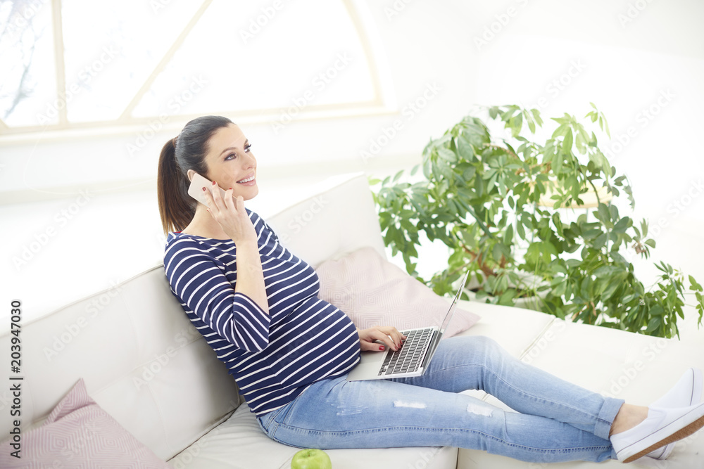 Obraz premium Pregnant woman talking on her mobile phone and using notebook while relaxing at home and sitting on couch
