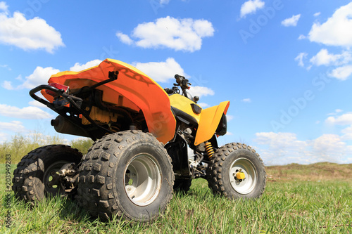 A four-wheeled yellow ATV quad-bike standing idle on the green grass, with trees and a blue sky with clouds on the background