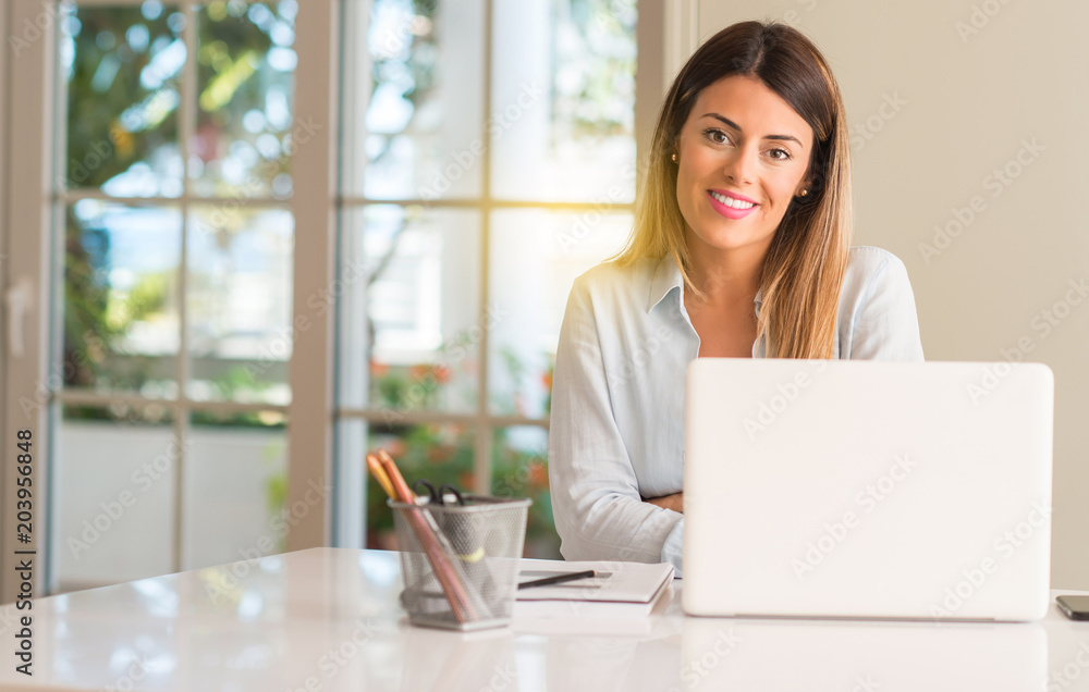 Student woman at table with laptop at home with crossed arms confident and happy with a big natural smile laughing Student woman at table with laptop at home with crossed arms confident and happy with a big natural smile laughing