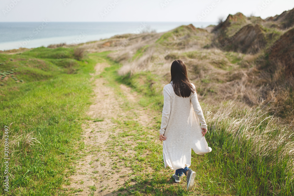 Back view Young brunette woman in light casual clothes walking along path in sunny weather in field near water on green background. Beautiful landscape. Lifestyle, leisure concept. Advertising area.