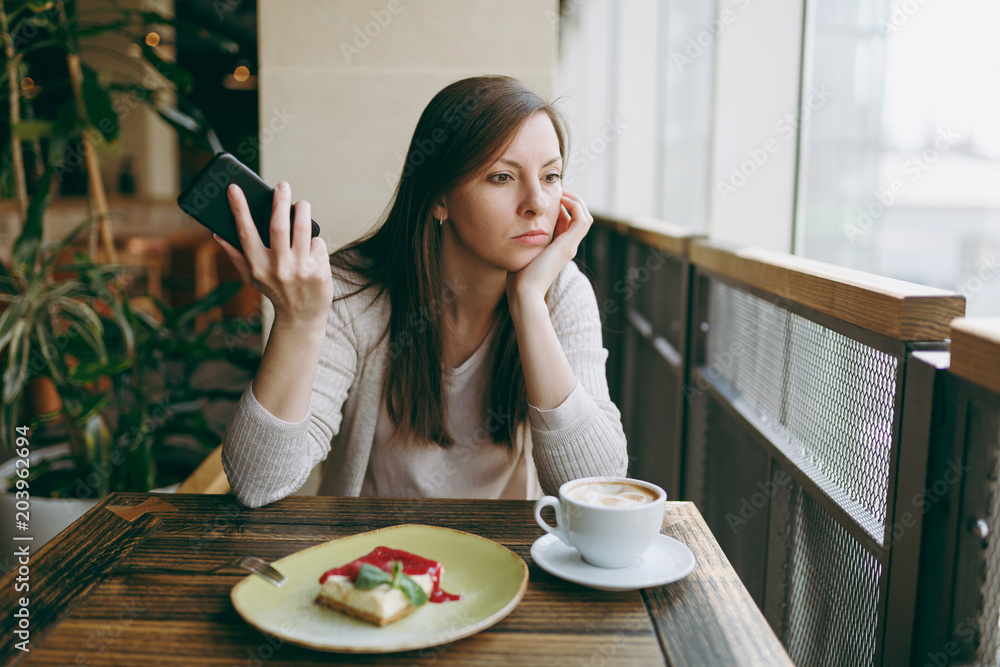 Young woman sitting alone in coffee shop at table with cup of cappuccino, cake, relaxing in restaurant during free time. Young female talking on mobile phone, having rest in cafe. Lifestyle concept.