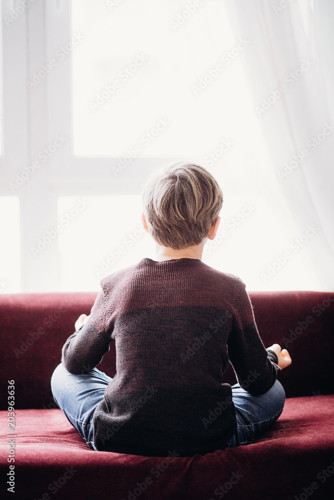a portrait of a boy sitting on a sofa in a lotus pose, view from his back, against the light window, soft focus a portrait of a boy sitting on a sofa in a lotus pose, view from his back, against the light window, soft focus