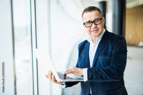 Professional at work. Confident senior man in suit and eyeglasses working on laptop while standing in office
