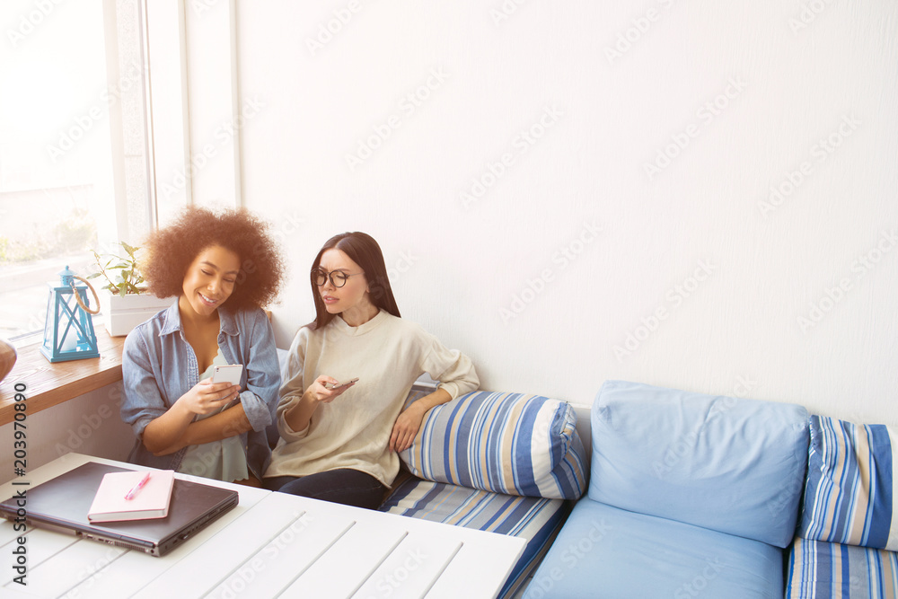 Nice picture of students sitting close to each other on sofa. THey are holding phones in their hands. Afro american girl is smiling while her friends is not happy and wondering. They have a break now. Nice picture of students sitting close to each other on sofa. THey are holding phones in their hands. Afro american girl is smiling while her friends is not happy and wondering. They have a break now.