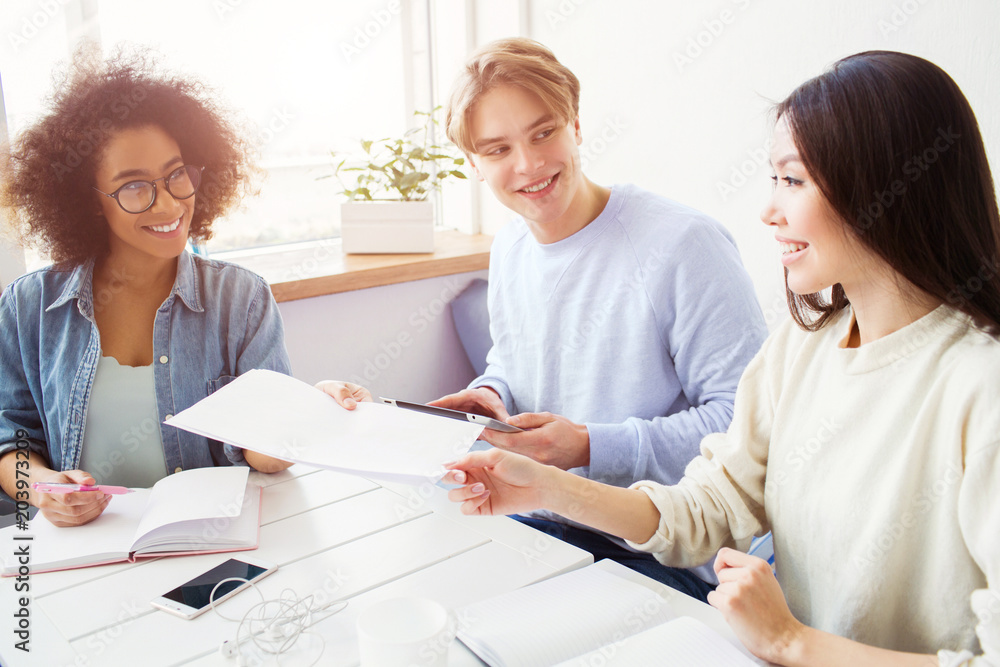 Nce picture of afro american and asian girls holding one piece of paper. There is also a guy sitting on sofa with girls and holding a tablet. He is looking and smiling to asian girl. Nce picture of afro american and asian girls holding one piece of paper. There is also a guy sitting on sofa with girls and holding a tablet. He is looking and smiling to asian girl.