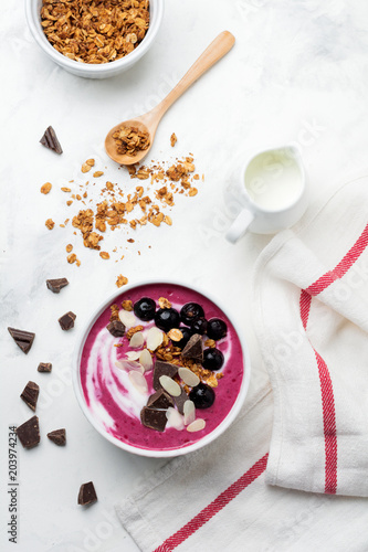 Pink black currant berries, banana smoothie bowl with granola, flakes from almonds and chocolate on light gray concrete background. Selective focus. Top view. Copy space.
