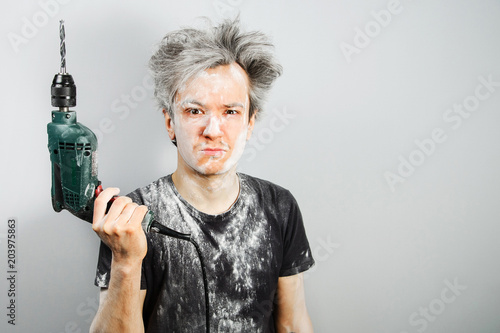 Fényképezés Unshorn and unshaven young guy soiled in plaster, holded drill and on gray background