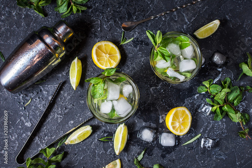 two glass of mojito cocktail with fresh lime and mint on a black stone board and bar shaker.