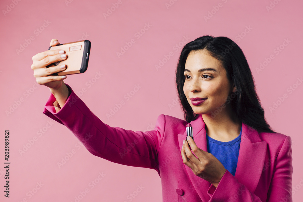 Φωτογραφία Portrait of a young woman in pink suit putting on lipstick using phone as mirror