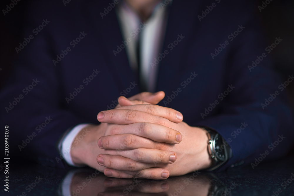 Obraz premium Businessman in blue suit and white shirt with crossed fingers sitting by the table on dark background