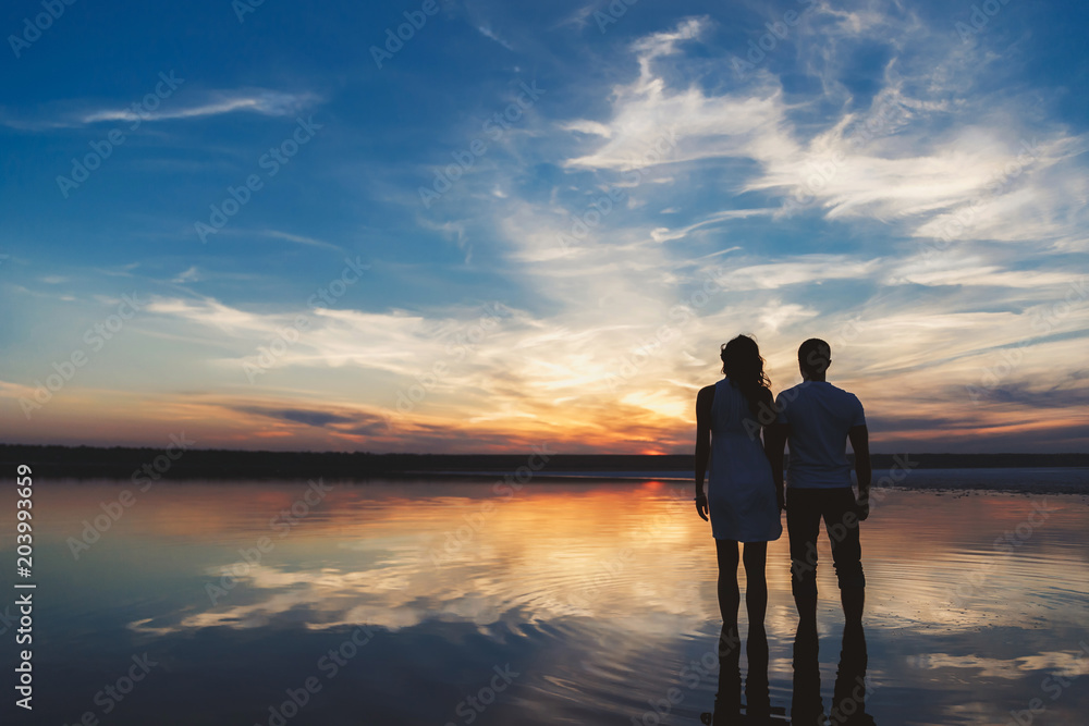 Portrait, silhouette of happy couple watching the colourful bright sunset standing in large lake, reflection in the water, holding hands