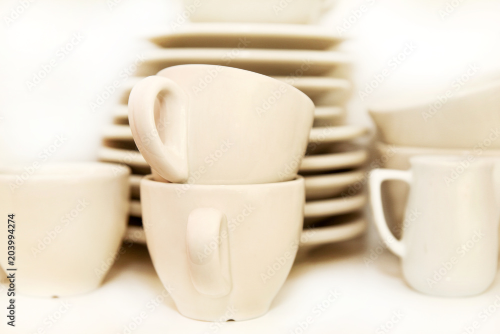 Small jug and white cups of coffee with service plate set on shelf on white background. Coffee cups with trays at the coffee bar. White crockery set. Cups, coffee, espresso and tea. Hot beverage. Small jug and white cups of coffee with service plate set on shelf on white background. Coffee cups with trays at the coffee bar. White crockery set. Cups, coffee, espresso and tea. Hot beverage.