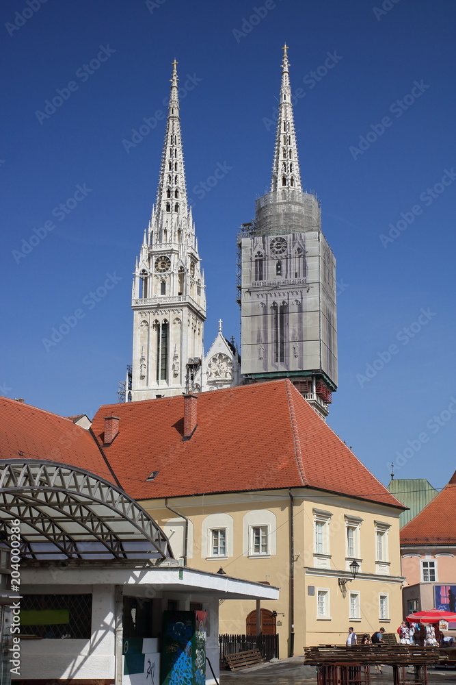 Centrum Zagrzebia, stolicy Chorwacji, z budynkiem zadaszonym pasażem oraz widocznymi zza budynku dwiema wieżami katedry katolickiej, jedna z wież restaurowana, z rusztowaniem, błękitne niebo - obrazy, fototapety, plakaty 