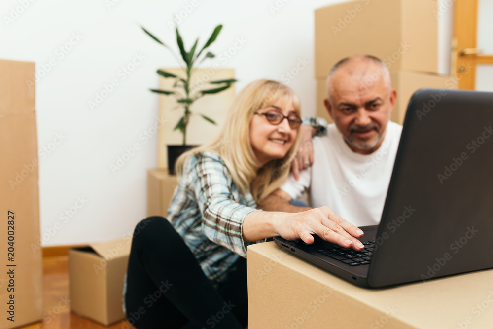 Middle aged couple sitting between boxes in their new apartment and using a laptop for some interior decorating ideas. Real estate theme. Middle aged couple sitting between boxes in their new apartment and using a laptop for some interior decorating ideas. Real estate theme.