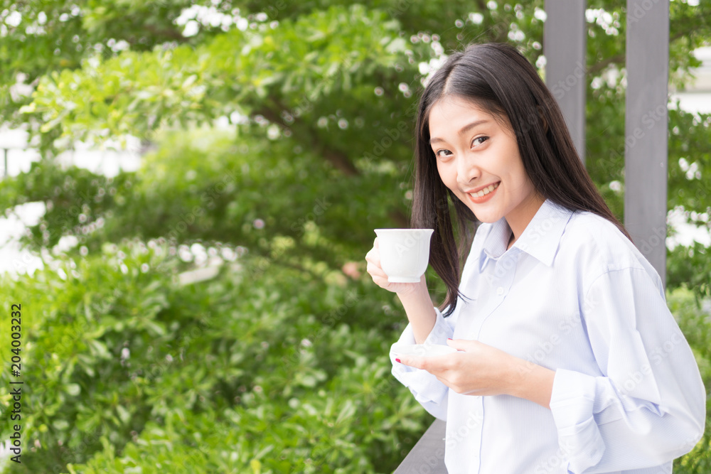 Beautiful young woman is holding cup of coffee looking at camera in the terrace with tree background. Beautiful young woman is holding cup of coffee looking at camera in the terrace with tree background.