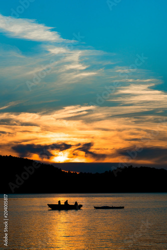 Sunsets through the clouds, while dad fishes with children (in silhouette) from a boat on the lake, while towing a kayak.