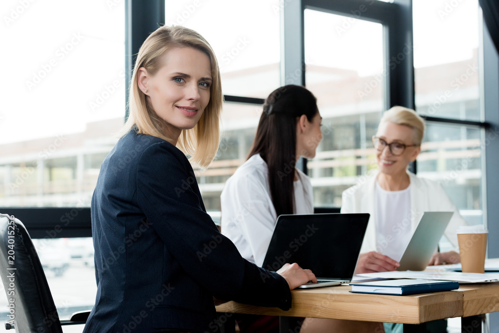 beautiful blonde businesswoman looking at camera at meeting while colleagues talking in office beautiful blonde businesswoman looking at camera at meeting while colleagues talking in office