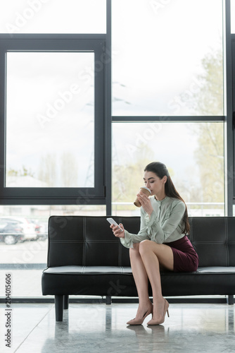 beautiful businesswoman holding smartphone and drinking coffee to go in office