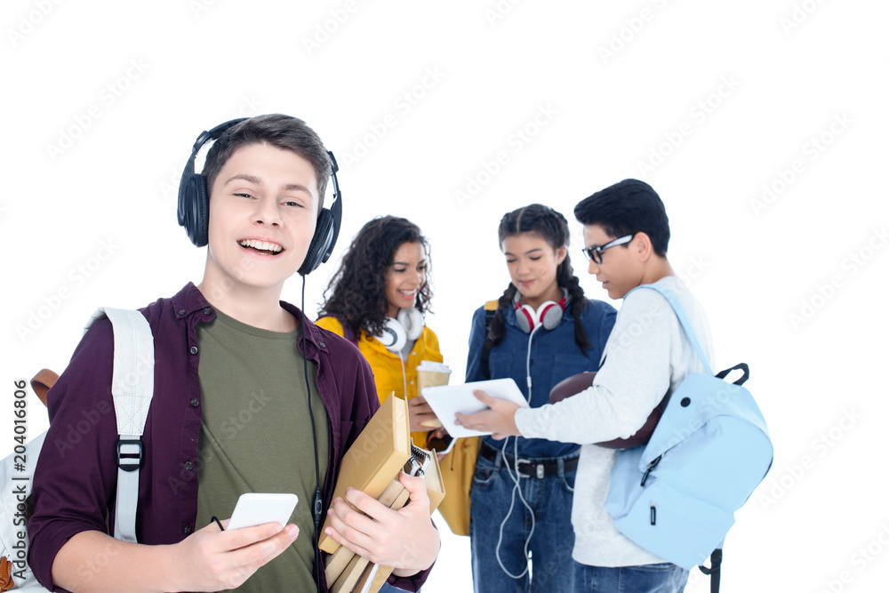 handsome teen student in headphones and with smartphone isolated on white with his friends chatting on background