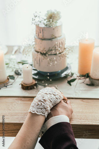 partial view of newlyweds holding hands while sitting at served table with wedding cake, rustic wedding concept