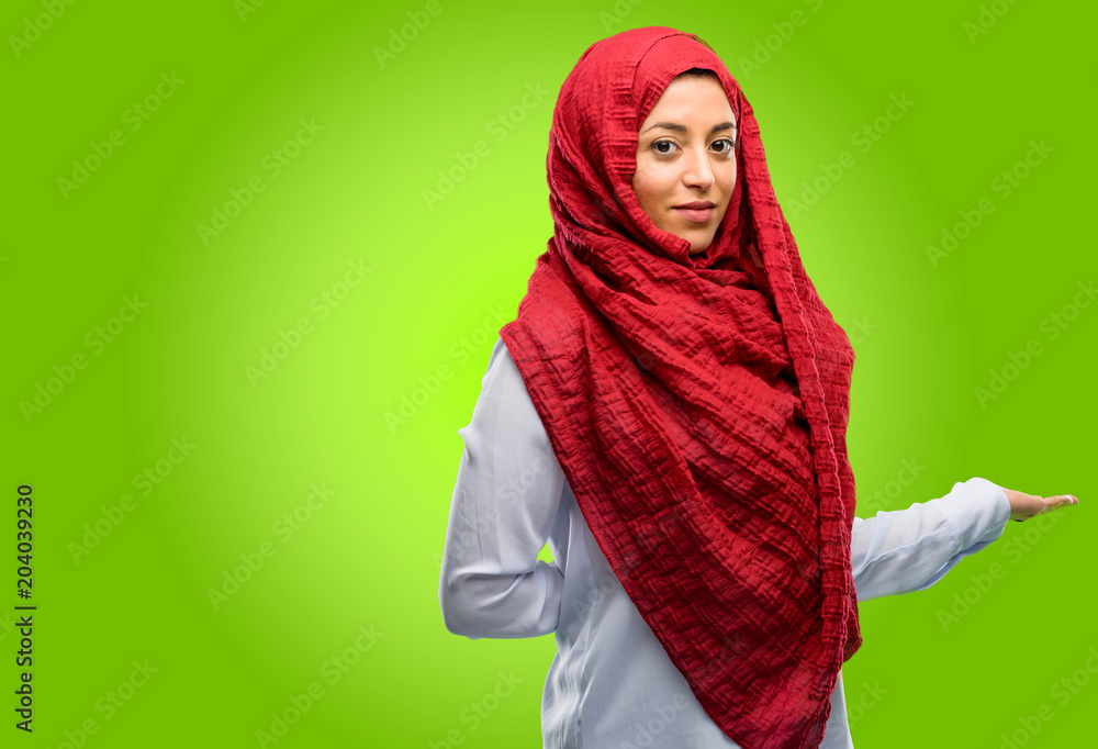 Young arab woman wearing hijab confident and happy with a big natural smile inviting to enter Young arab woman wearing hijab confident and happy with a big natural smile inviting to enter