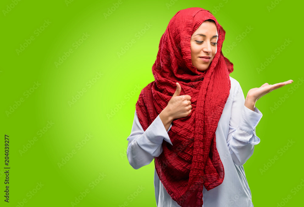 Young arab woman wearing hijab thumb up holding something in his empty hand and making ok gesture Young arab woman wearing hijab thumb up holding something in his empty hand and making ok gesture