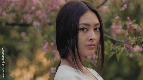 Gorgeous asian woman with perfect skin creartive art make-up wearing trendy white japanese kimono standing agaist flower background in the garden and looking at camera. Close-up fashion portrait photo