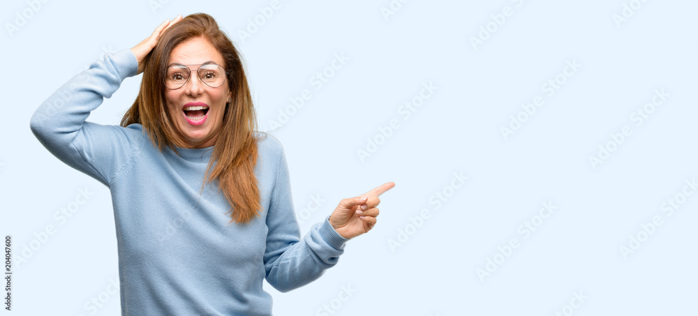 Middle age woman wearing wool sweater and cool glasses pointing away side with finger isolated blue background Middle age woman wearing wool sweater and cool glasses pointing away side with finger isolated blue background