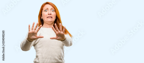 Beautiful young redhead woman disgusted and angry, keeping hands in stop gesture, as a defense, shouting isolated over blue background