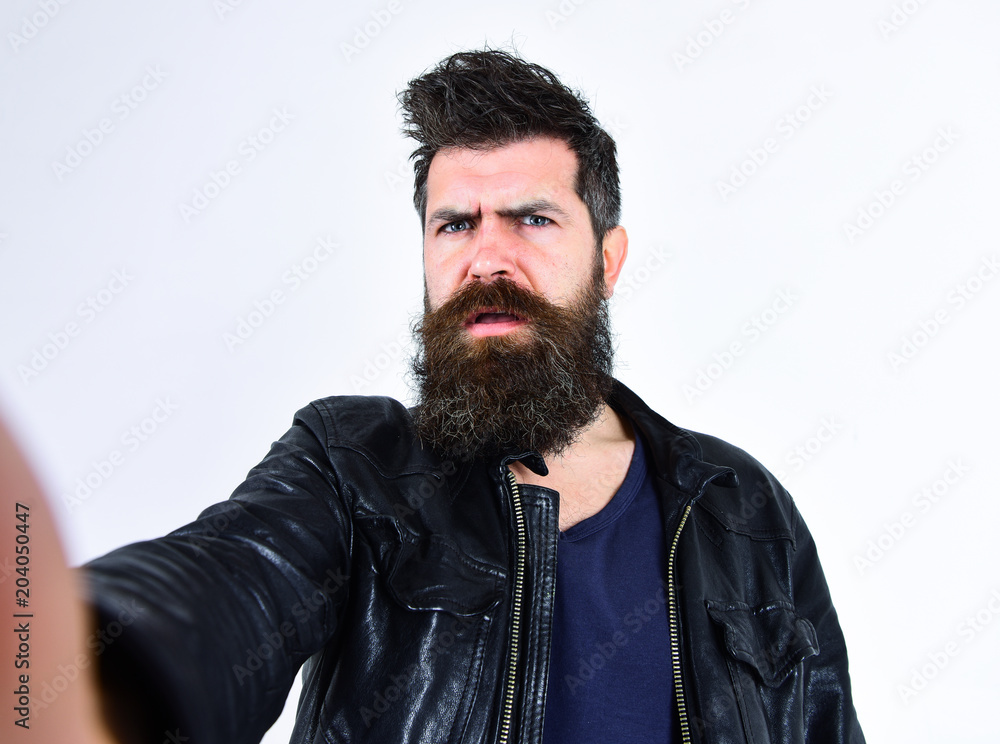Man with beard and mustache on strict face looking at camera. Macho wears leather jacket, white background. Hipster looks serious and thoughtful while taking selfie photo. Masculinity concept. Man with beard and mustache on strict face looking at camera. Macho wears leather jacket, white background. Hipster looks serious and thoughtful while taking selfie photo. Masculinity concept.