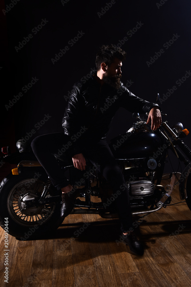 Man with beard, biker in leather jacket lean on motor bike in darkness, black background. Macho, brutal biker in leather jacket stand near motorcycle at night time, copy space. Biker culture concept.