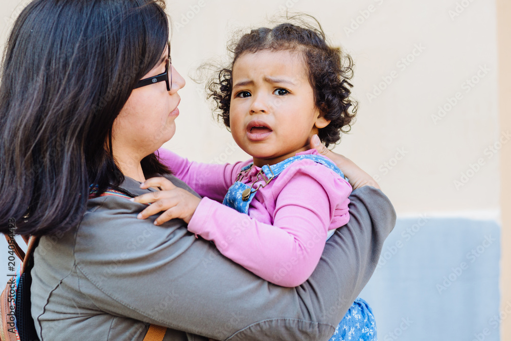 Close up of unhappy dark skinned toddler baby girl crying while her unrecognizable ecuadorian mother trying to calming down and holding her on hands. No to violence against children concept. Close up of unhappy dark skinned toddler baby girl crying while her unrecognizable ecuadorian mother trying to calming down and holding her on hands. No to violence against children concept.