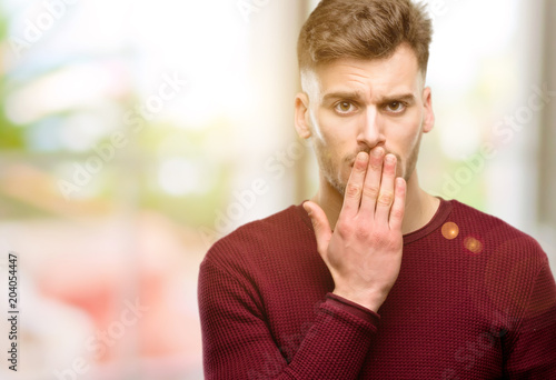 Handsome young man covers mouth in shock, looks shy, expressing silence and mistake concepts, scared