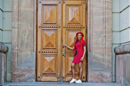 Cute and slim african american girl in red dress with dreadlocks and backpack posed against school large wooden door. Stylish black student.