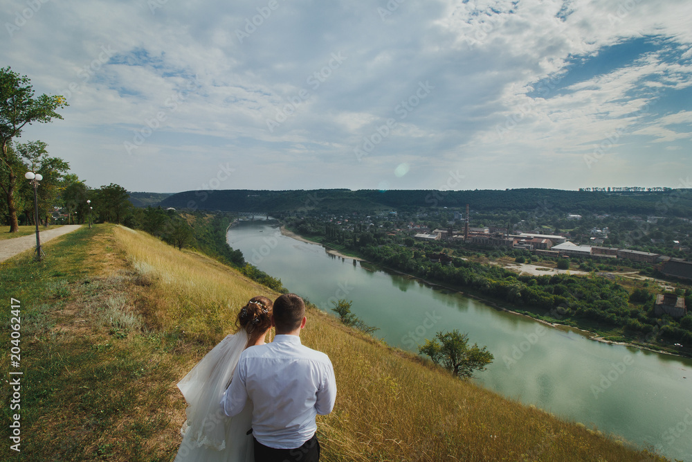 Wedding couple is hugging looking at the beautiful view on the river and village in summer. Amazing natural landscape with dry yellow grass and green trees. Bride in puffy dress and groom embrace.