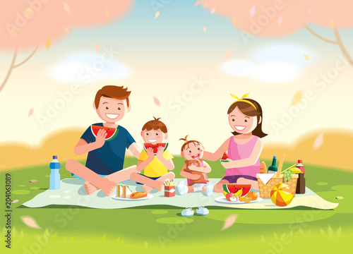 Family enjoying picnic. They are sitting and eating watermelon on the grass in a park, the basket with meal and toys for the kids. Blurred background. 