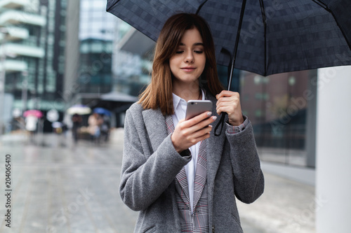 Attractive female manager using mobile phone in modern area, young girl browsing phone smiling under umbrella while it is raining outdoors, technology, smartphone and wireless connection concept