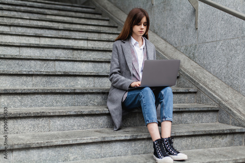Young stylish fashion blogger using laptop for work while sitting outside on staircase, beautiful busy woman typing on portable computer for distance job, businesswoman working on pc in the city © makarovada