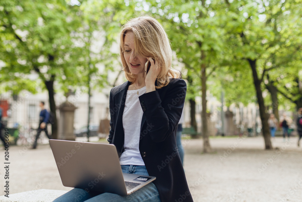 Beautiful female manager typing on portable computer for distance job and talking with her cell phone sitting in a park during a break, businesswoman working on pc in the fresh air Beautiful female manager typing on portable computer for distance job and talking with her cell phone sitting in a park during a break, businesswoman working on pc in the fresh air
