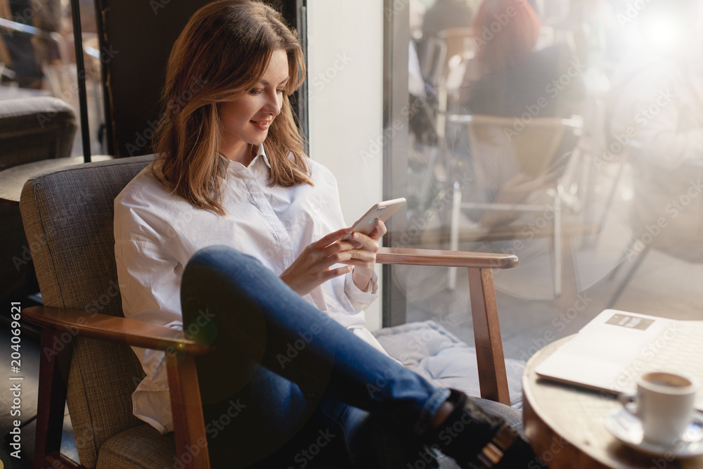 Happy young businesswoman using mobile phone in coffee shop, beautiful female freelancer in casual style chatting in smatrphone sitting relaxed at cafe near the window in armchair and having a break