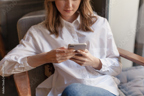 Happy young businesswoman using mobile phone in coffee shop, cropped shot of beautiful female freelancer in casual style chatting in smatrphone sitting relaxed at cafe in armchair and having a break