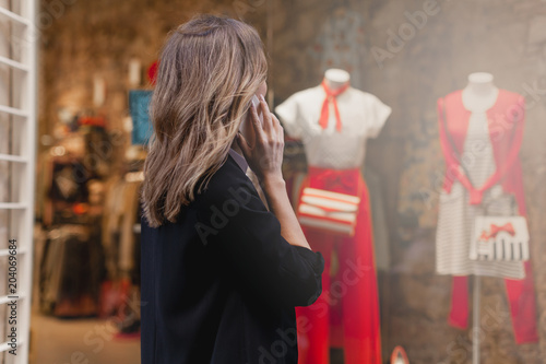 Young beautiful woman walking along the street talking via mobile phone and doing shopping, stylish girl looking at clothes shop window while having a conversation with friend on smartphone