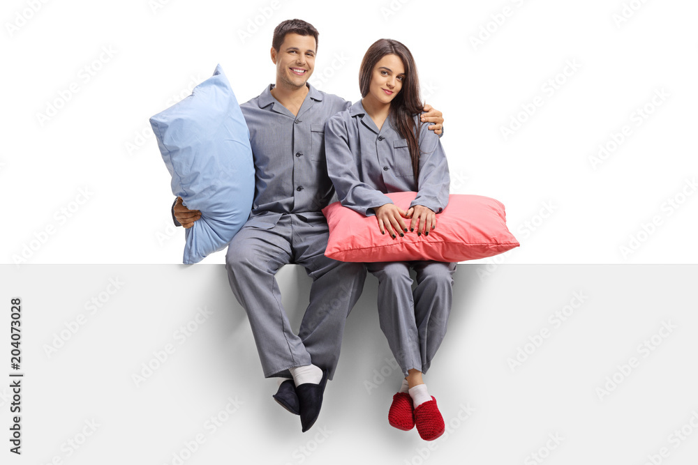 Young couple dressed in pajamas sitting on a panel and holding pillows - obrazy, fototapety, plakaty 