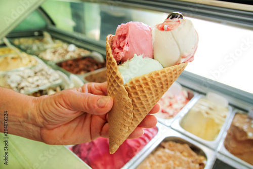 Female hand is holding a large strawberry, mint, vanilla with sour cherry ice cream in waffle cone. Ice cream fridge with steel service containers in background. Worker in ice cream shop. Pastry shop.