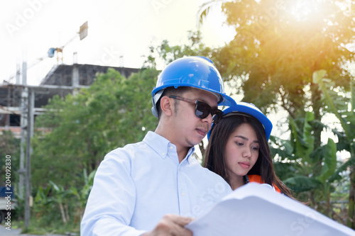 Asian man and woman engineer or architect with the blue safety helmet meeting at the construction site with blueprints and laptop. © Thaspol