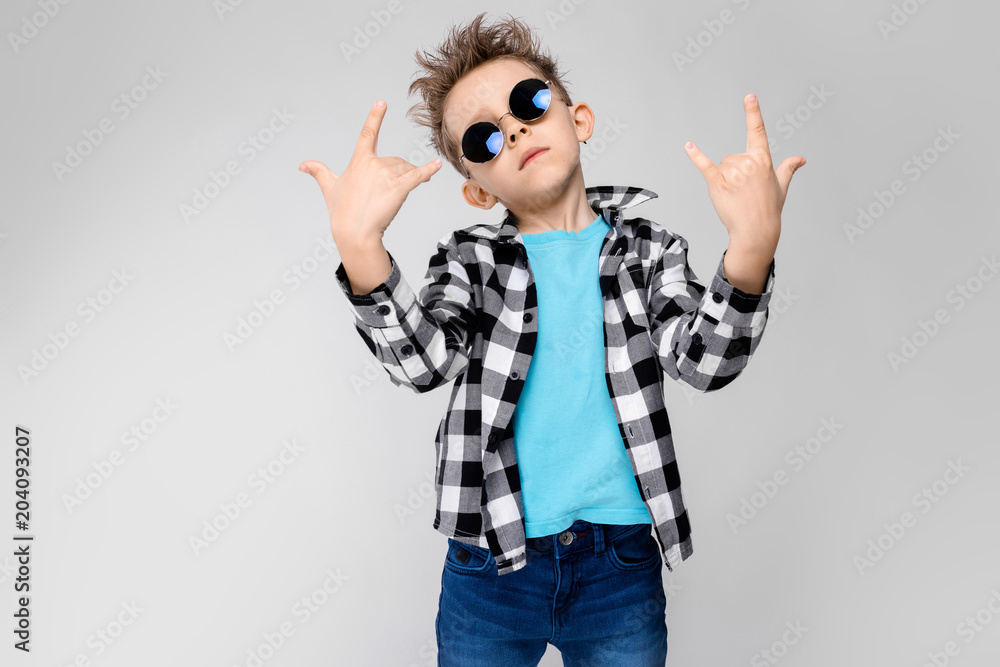 blue shirt and jeans stands on a gray background. The boy is wearing round glasses. Redhead boy shows a rocker goat blue shirt and jeans stands on a gray background. The boy is wearing round glasses. Redhead boy shows a rocker goat