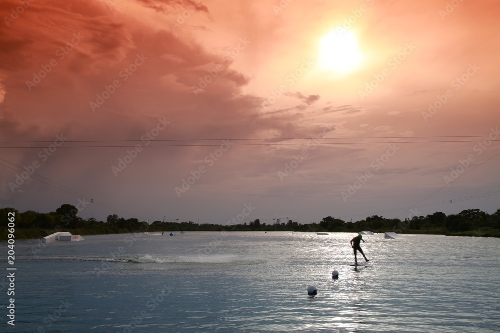 Fototapeta premium Wakeboarder Doing Half-Flip Being Pulled by Cable on Lake Against Late Afternoon Sun through Orange Graduated Filter