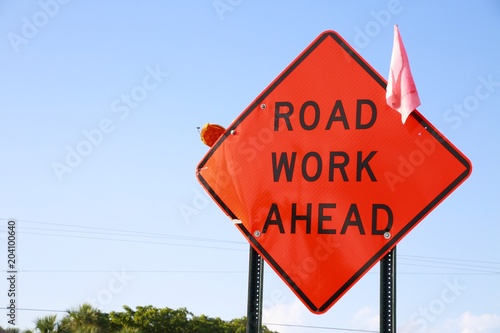 Orange Road Work Ahead Diamond Shaped Sign with Tiny Pink Flag, Clear Blue Sky and Trees Behind