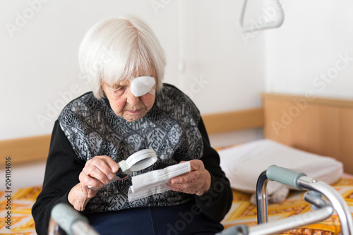 Visually impaired elderly 95 years old woman sitting at the bad trying to read her medical therapy prescription with magnifying glass due to eyesight problems. photo